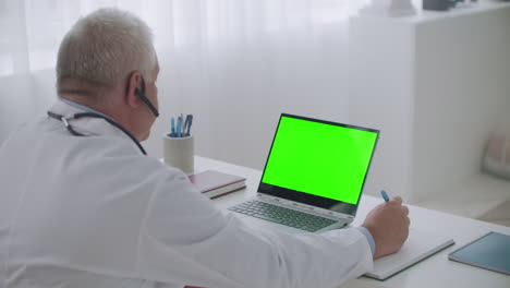Ein-Männlicher-Arzt-Berät-Sich-Online-Von-Seinem-Büro-Aus-Auf-Einem-Laptop-Mit-Grünem-Display-Für-Chroma-Key-Technologie-Und-Blickt-Auf-Den-Bildschirm
